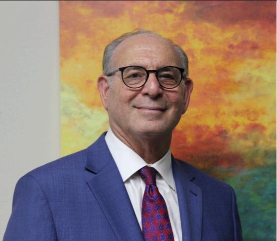 A man in a blue suit and a red and blue tie is standing and smiling in front of a painting